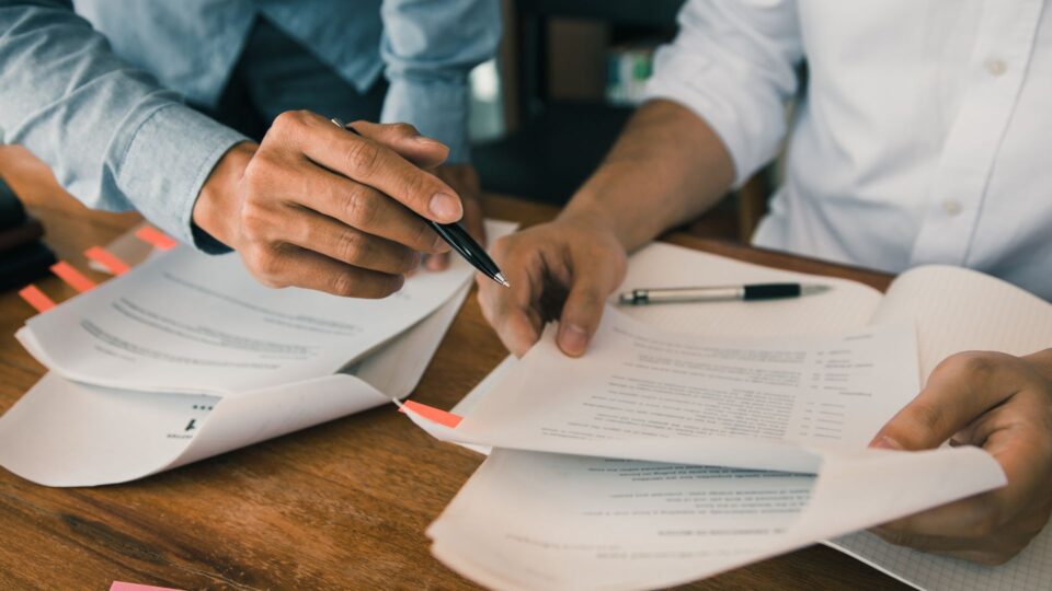This image captures a close-up of two individuals' hands engaged in a professional or collaborative task involving documents and a pen.